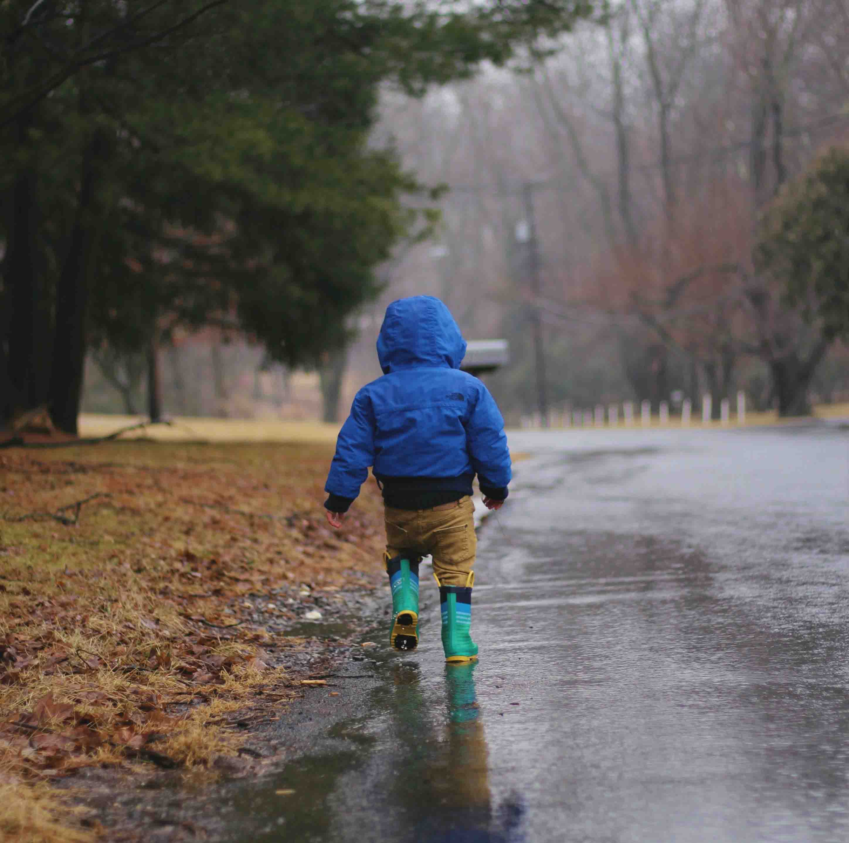 A toddler in a blue jacket and green and yellow wellies stomps through the puddles.